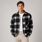 A man modeling a long-sleeve plaid flannel shirt over a white t-shirt and beige pants, standing against a light gray studio background.