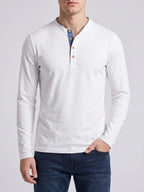 Model wearing a Blue Slim-Fit cotton long-sleeve Henley shirt on a sunlit garden patio beside a stone house.
